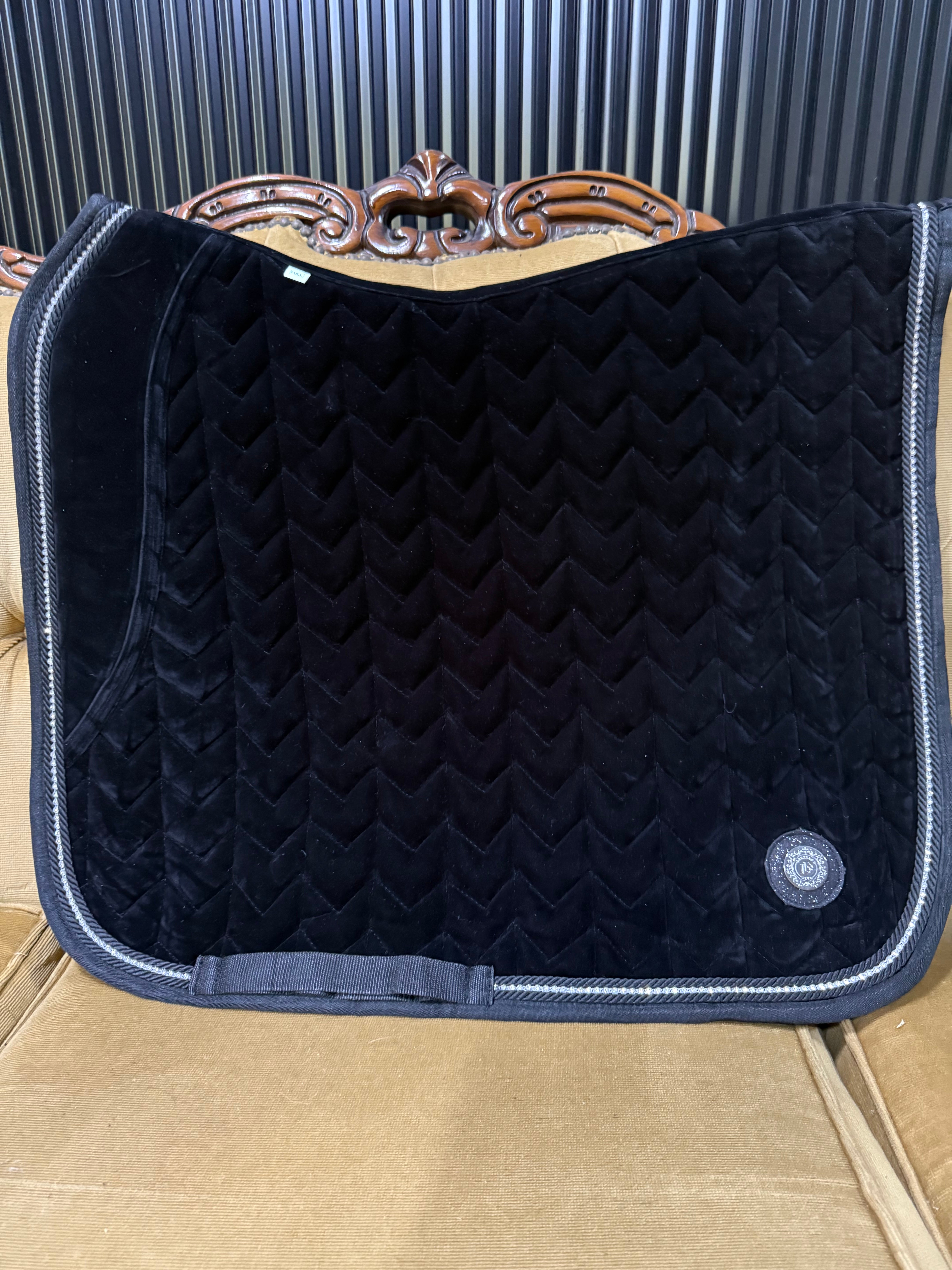 Black velvet saddle pad with a single row of crystal trim and a signature metal logo on the lower corner, displayed on a saddle.